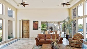 A spacious living room with a brown leather sofa, a patterned armchair, and a coffee table with a vase. There are two ceiling fans, a wooden door, and large windows offering views of the outdoors. - Red Cliffs (Washington)