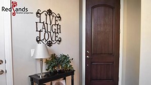 A dark wooden door, a wall-mounted decorative sign saying "LIVE LAUGH LOVE", a table with a lamp and a plant, and a framed picture on the table. - Solana Linda (Washington)