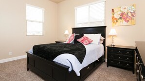 Un dormitorio con una cama negra, almohadas blancas y rojas, una mesita de noche con una lámpara, un aparador y un cuadro floral en la pared.