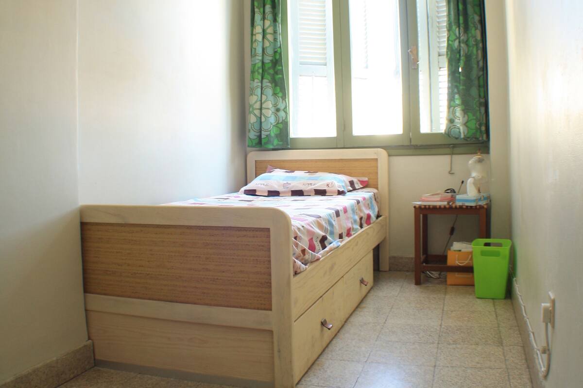 A small bedroom with a single bed, a window with curtains, a bedside table, and a green trash bin.