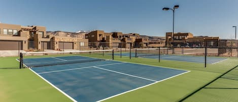 A tennis court with a net, surrounded by a fence and buildings in the background.