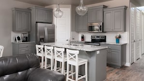 A modern kitchen with grey cabinets, a stainless steel refrigerator, a microwave, and a central island with bar stools. - Your Escape at the Ledges (St. George)