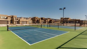 Un court de tennis avec un filet, entouré d’une clôture et avec des bâtiments en arrière-plan.