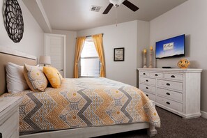 Une chambre à coucher comprenant un lit, une commode, un ventilateur de plafond, une fenêtre avec des rideaux et un téléviseur fixé au mur.
