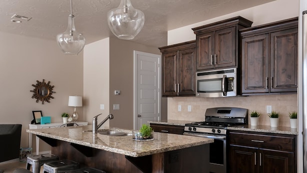 A modern kitchen with dark wood cabinets, a granite countertop, stainless steel appliances, and a central island with stools. - The Perfect Place (St. George)