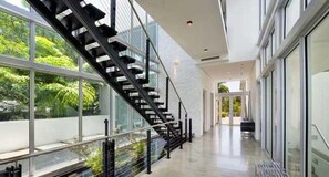 A modern interior with a glass staircase leading to a balcony, surrounded by large windows offering views of greenery. - Villa Angela (Miami Beach)