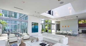 A modern living room with a large glass wall, white furniture, and a dining area with white chairs. - Villa Angela (Miami Beach)