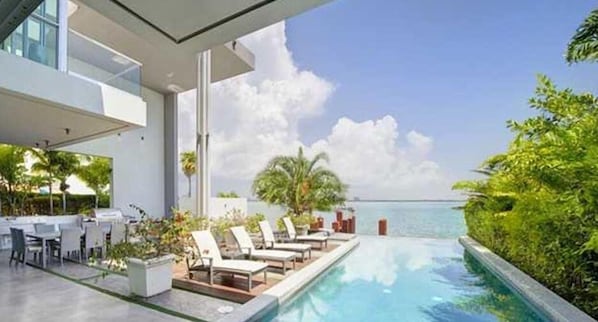 A modern pool area with lounge chairs and a dining setup overlooking a body of water. - Villa Angela (Miami Beach)