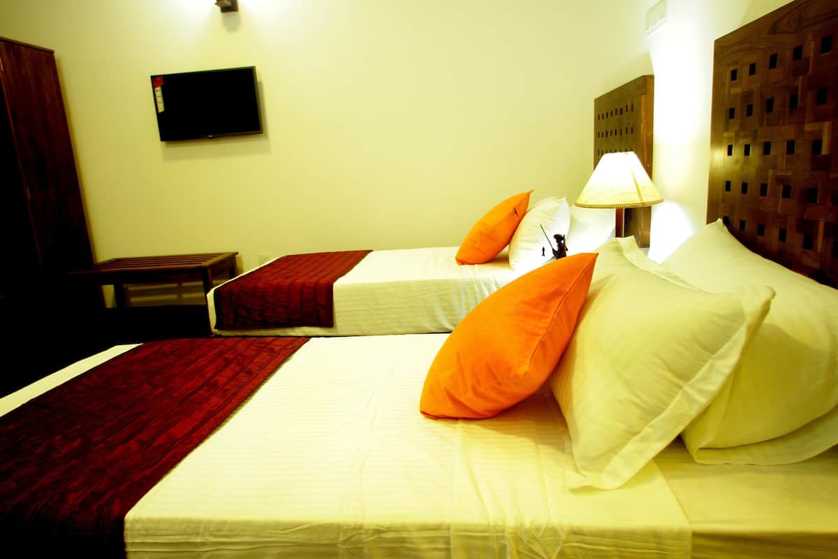 Une chambre d’hôtel avec deux lits, chacun agrémenté de draps blancs et d’oreillers orange. Un téléviseur est fixé au mur au-dessus des lits, et un chevet en bois se trouve sur le côté droit.