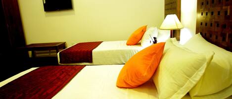 Quarto de hotel com duas camas, cada uma com roupa de cama branca e almofadas laranja. Há uma televisão montada na parede acima das camas e uma cabeceira de madeira no lado direito.