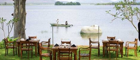 A lakeside dining area with wooden tables and chairs, a small boat on the water, and a distant view of mountains.