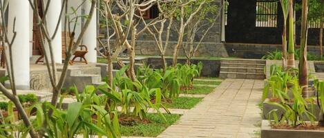 A pathway leading to a building with a thatched roof, surrounded by greenery and stone pillars.