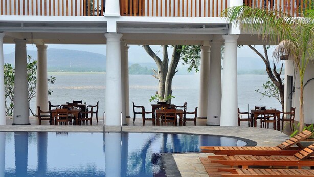 A poolside area with wooden lounge chairs and tables, a view of the water, and a covered seating area with columns. - Peacock Wild Yala (Thissamaharama)