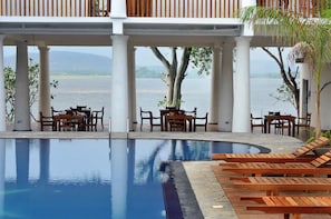 A poolside area with wooden lounge chairs and tables, a view of the water, and a covered seating area with columns.