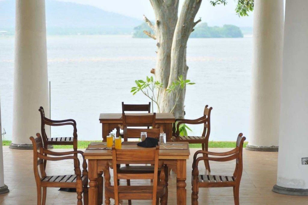 Een houten tafel met stoelen buiten, met uitzicht op een groot wateroppervlak en een boom op de achtergrond.