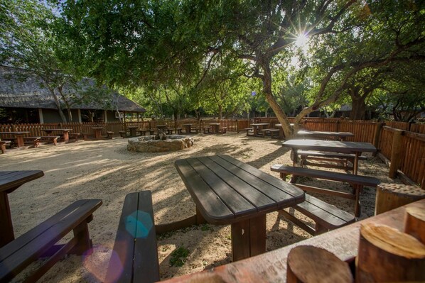An outdoor seating area with wooden picnic tables and benches, surrounded by trees and a thatched-roof structure.