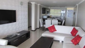 Una sala de estar moderna con una televisión de pantalla plana, un sofá con cojines rojos, una mesa de centro y un comedor con sillas blancas.