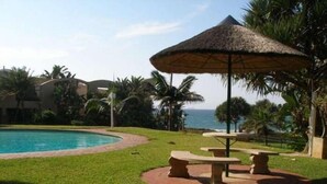Zona junto a la piscina con una sombrilla de paja, bancos de piedra y vistas al océano.