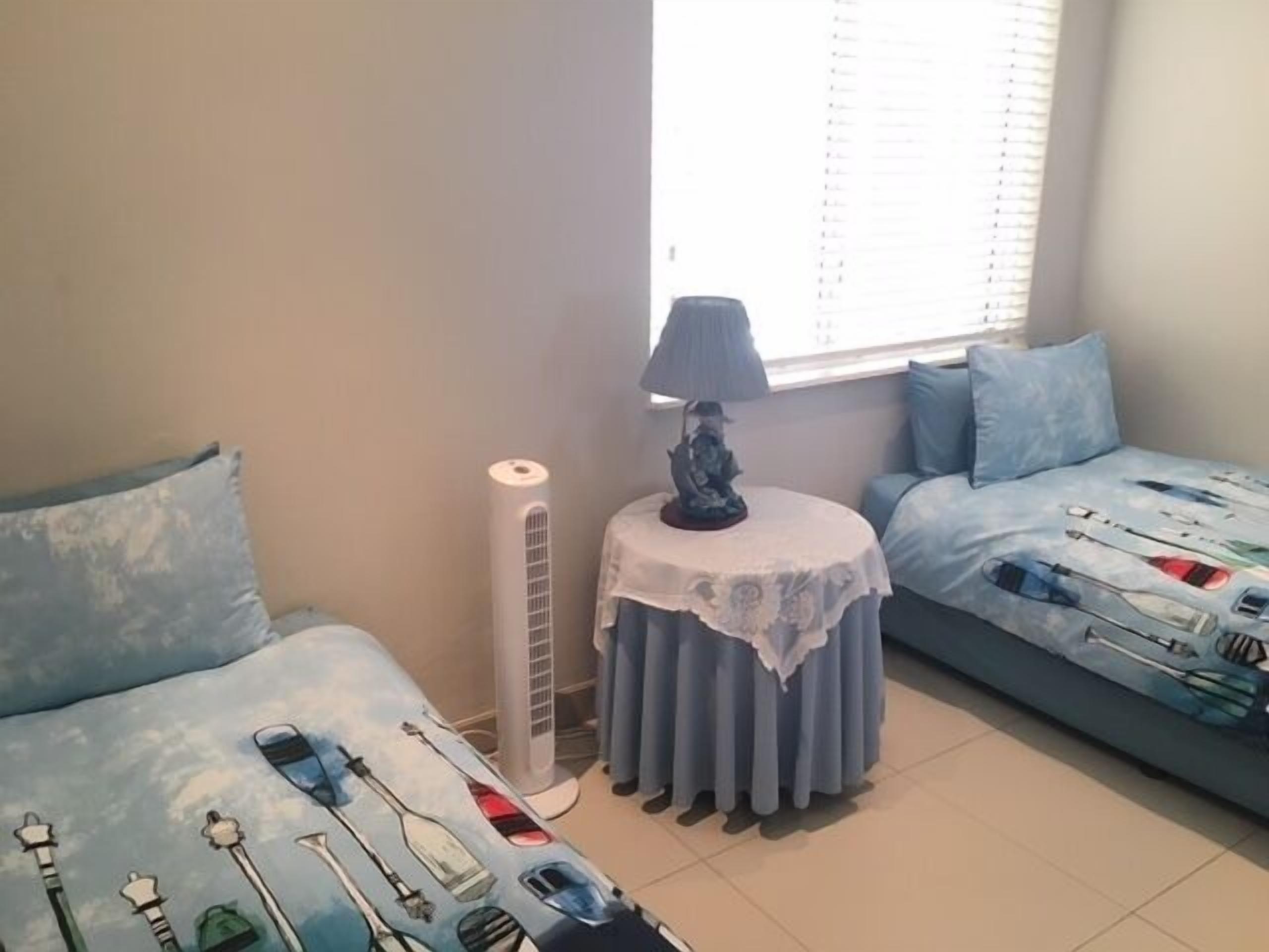 A bedroom with two beds, a standing heater, a table with a lamp, and a window with blinds.