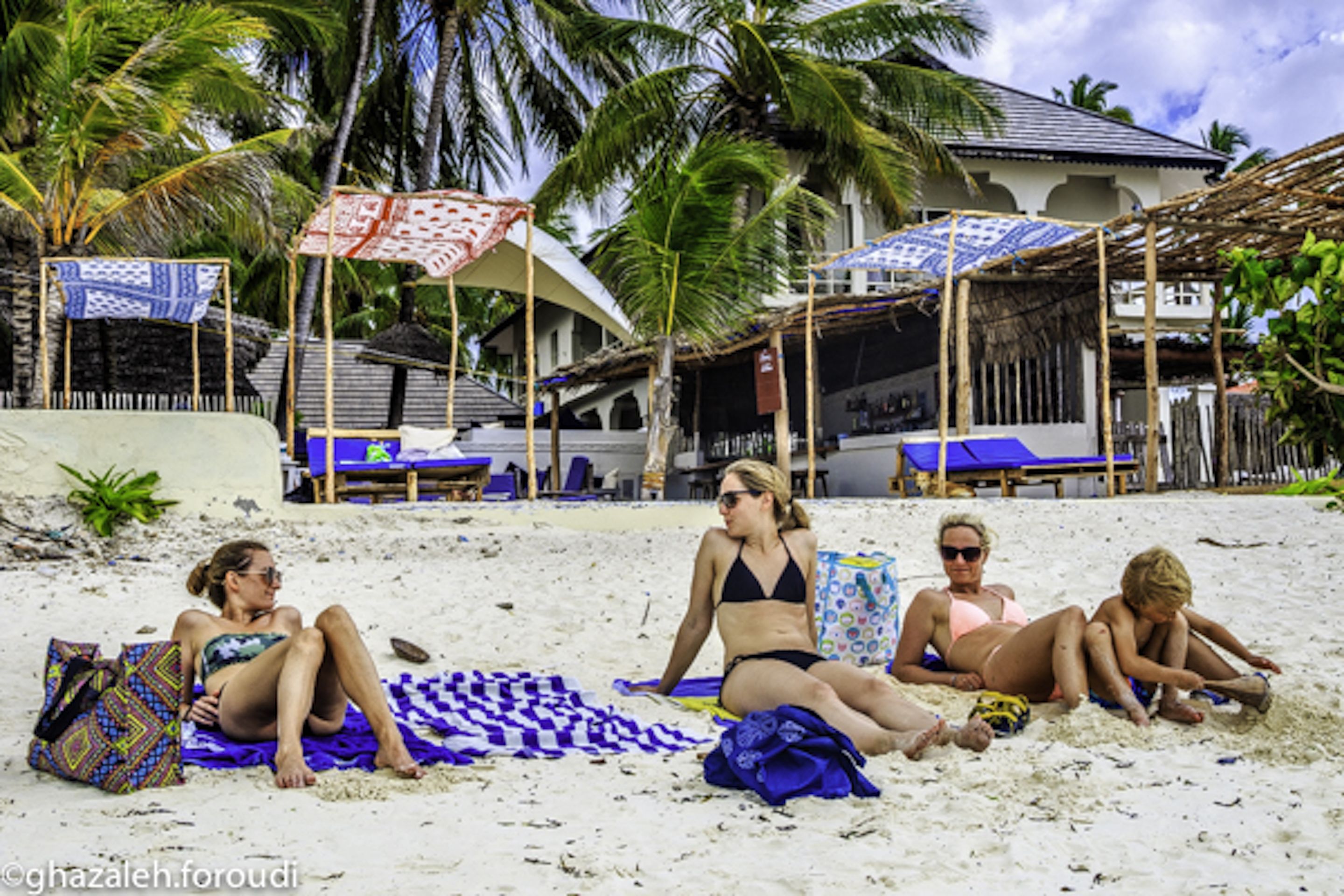 A beach scene with people relaxing on towels, umbrellas, and a building with a thatched roof in the background.
