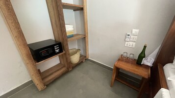 Un angolo di una stanza con un ripiano in legno che contiene un dispositivo elettronico nero, un cesto intrecciato e un piccolo tavolino con due bicchieri e una bottiglia.