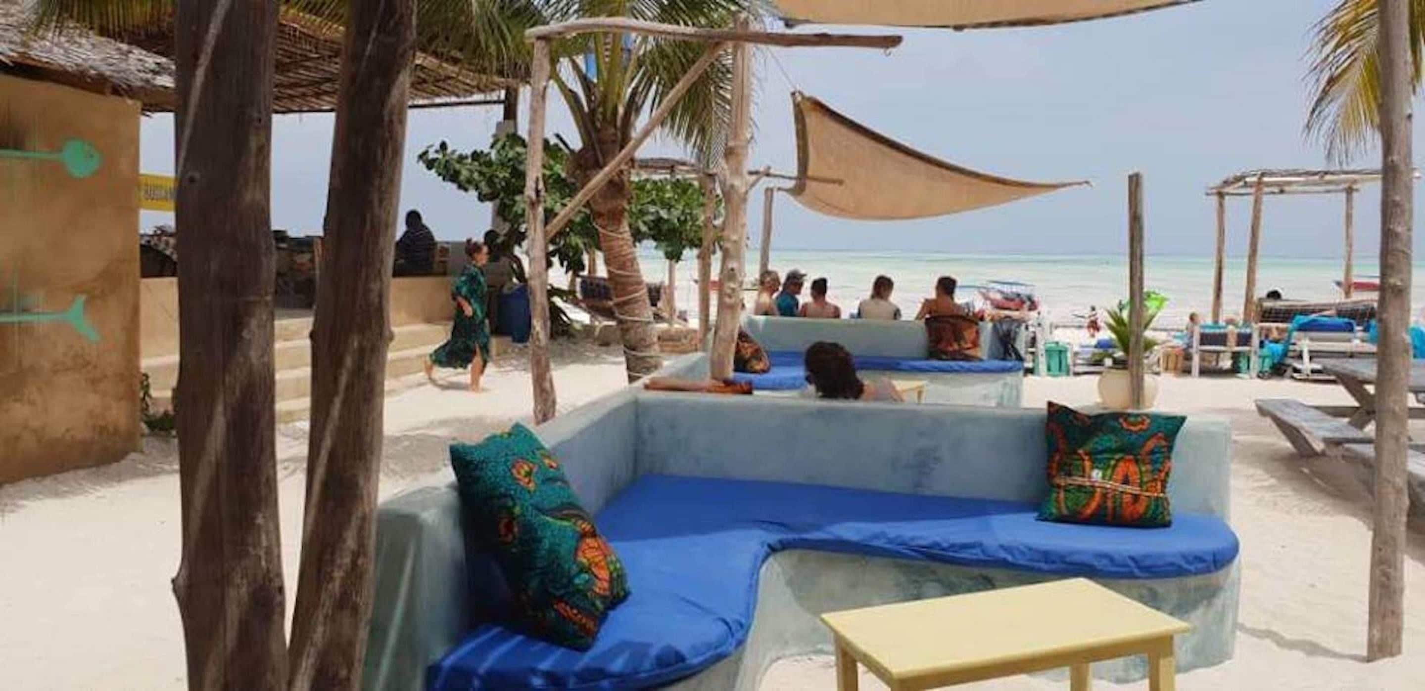 Un área de descanso en la playa con asientos acolchados azules y una pequeña mesa, con vista al océano.