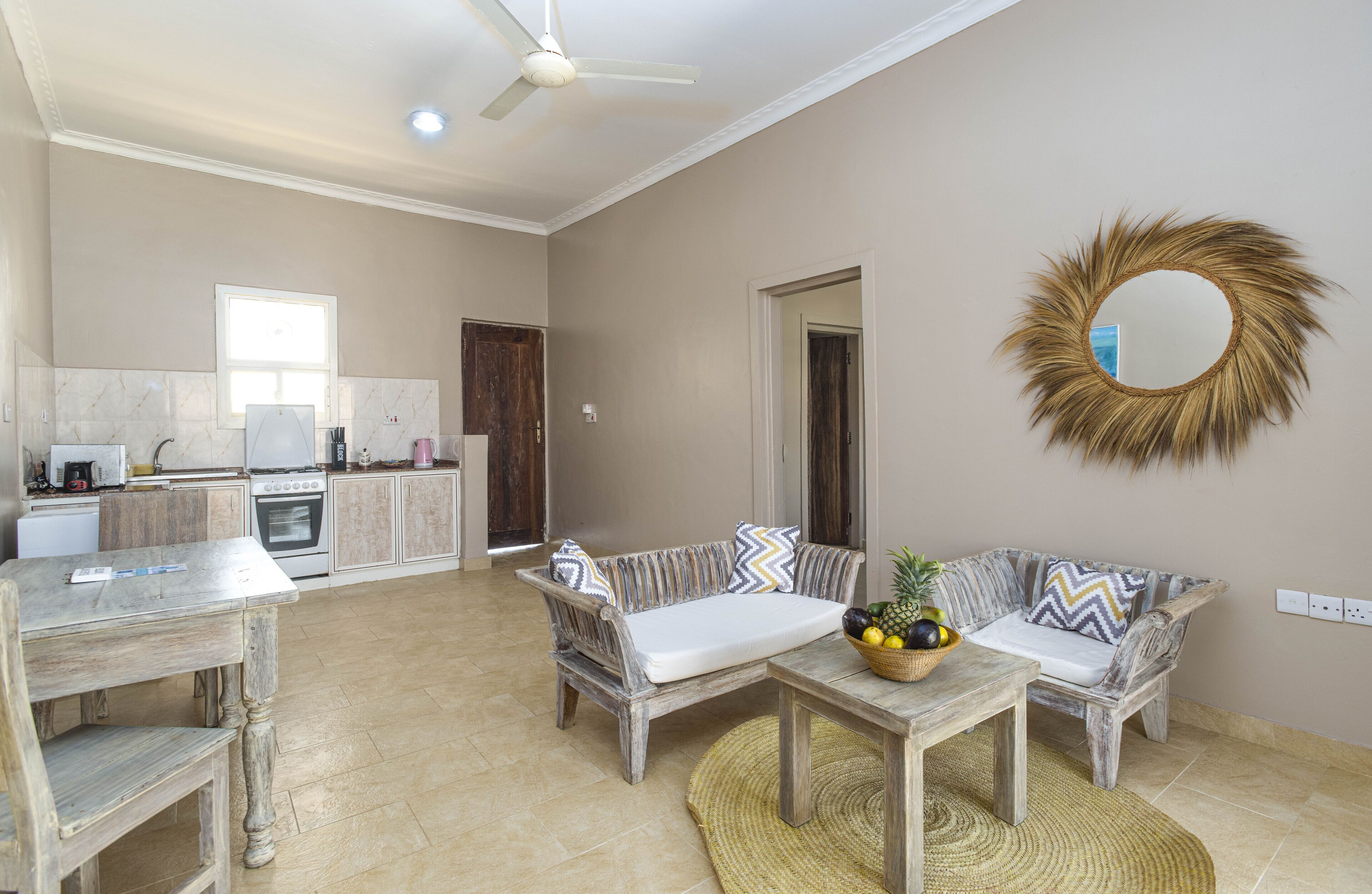 A spacious living area with a sofa set, a coffee table with fruit, a wooden table, and a kitchen in the background.