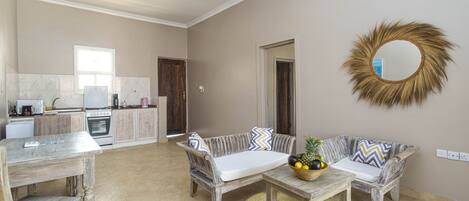 A spacious living area with a sofa set, a coffee table with fruit, a wooden table, and a kitchen in the background.