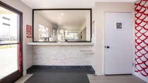 A hotel reception area with a white brick counter, a door with a red decorative panel, and a view of the street outside. - Hotel Palmdale - Antelope Valley (Palmdale)