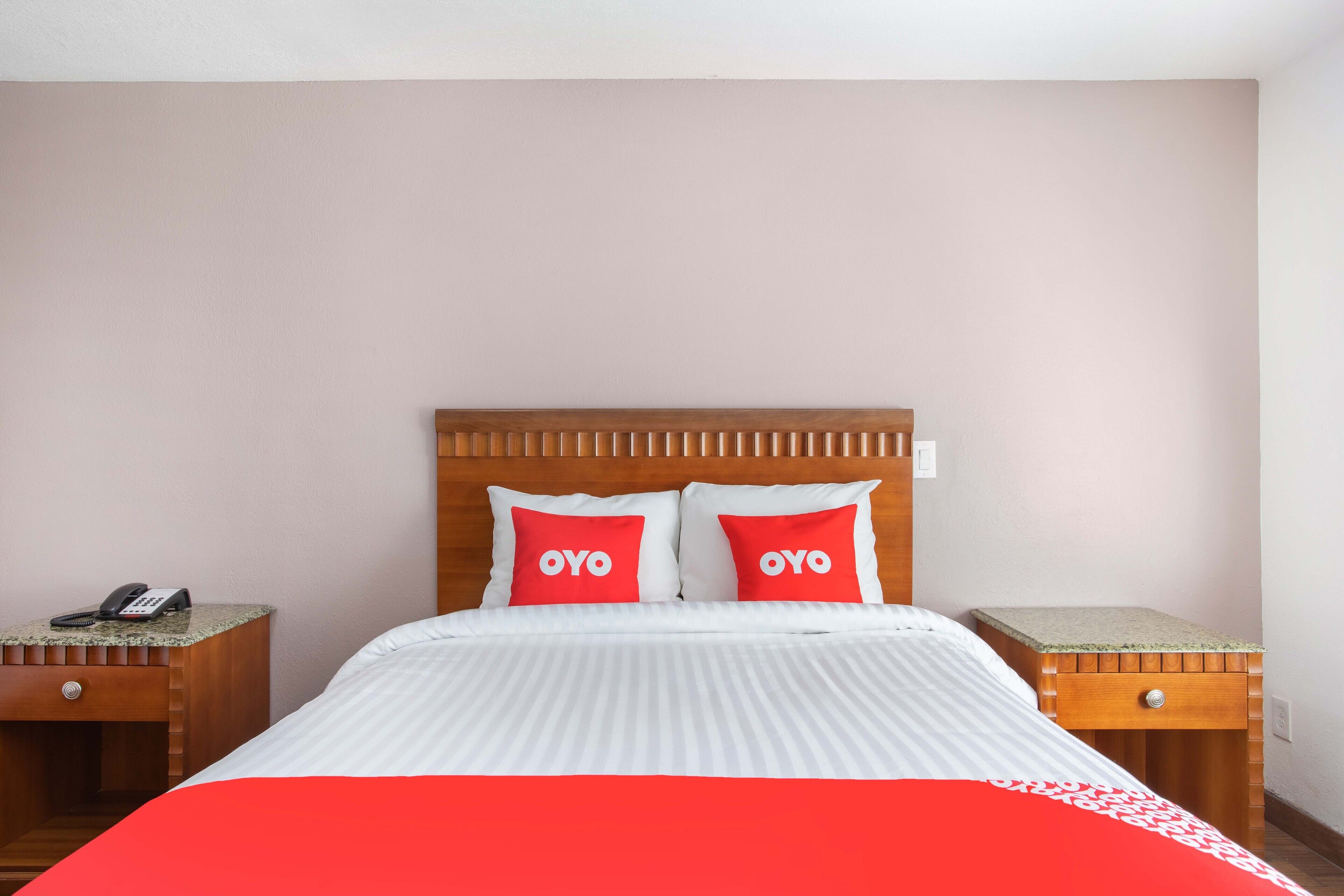 A hotel room with a bed, two pillows with the word "OYO" on them, a wooden headboard, a bedside table with a phone, and a small drawer.