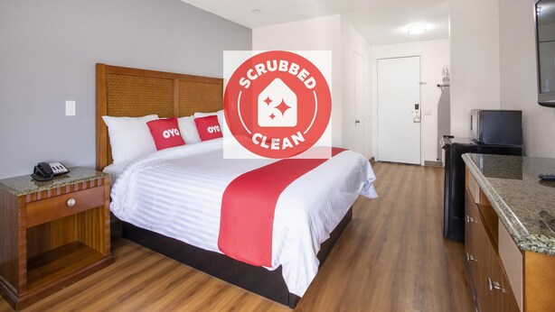 A hotel room with a bed, wooden headboard, a nightstand with a phone, a red and white bedspread, a red pillow, a TV, and a kitchen area with a microwave. - Hotel Palmdale - Antelope Valley (Palmdale)