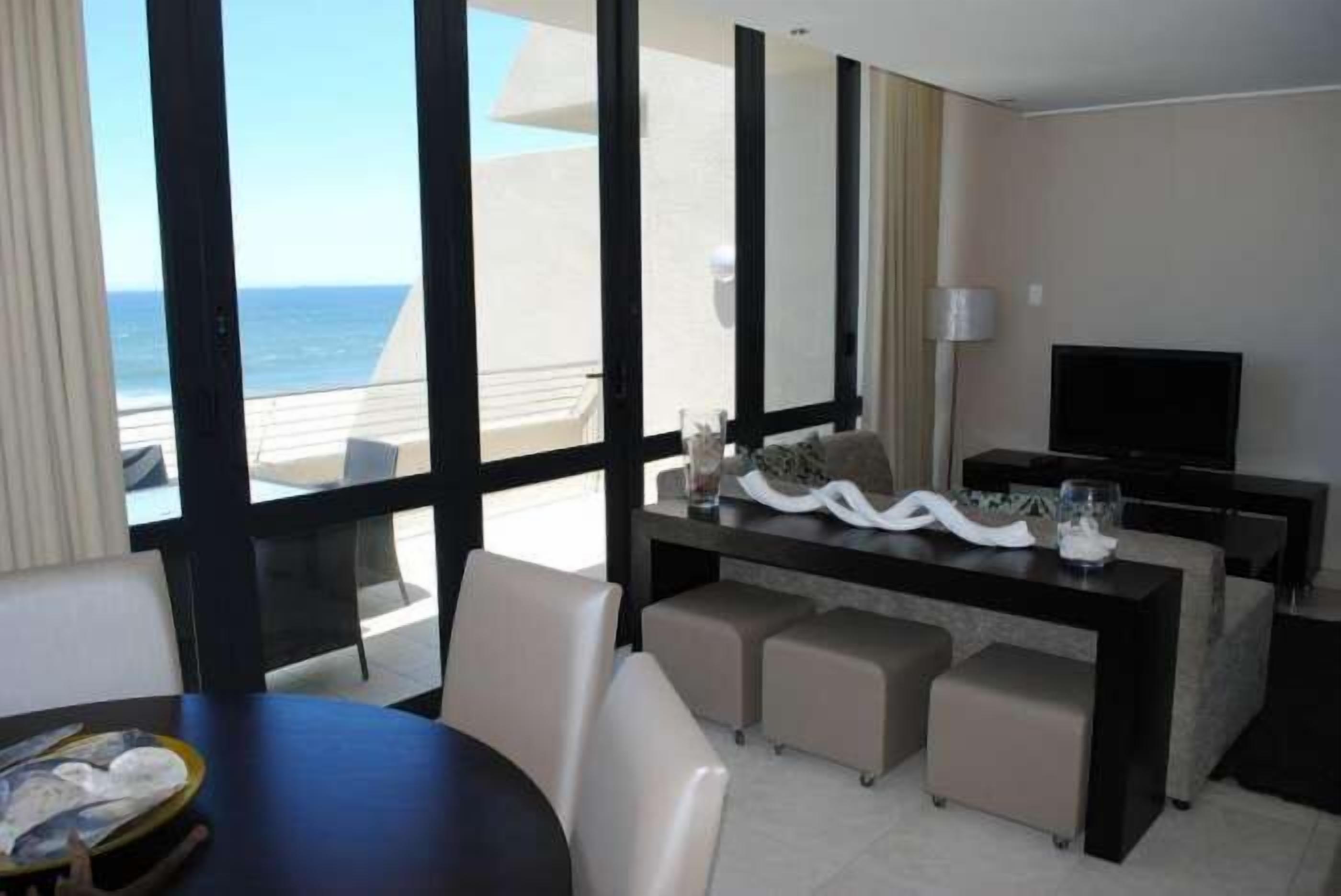 A hotel room with a balcony, a dining area with a table and chairs, a sofa, a TV, and a view of the ocean.