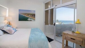 Un dormitorio con cama, un cuadro de una escena costera, una puerta corrediza de cristal a un balcón y una mesita de noche de madera con lámpara.