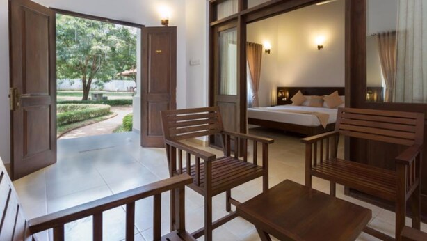 A hotel room with a bed, a desk, and two chairs. There is a door leading to a balcony with a view of trees and a pathway. - Chenra Hotel (Kataragama)