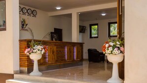 A reception area with a wooden reception desk, two potted plants with flowers, and a clock on the wall. - Chenra Hotel (Kataragama)