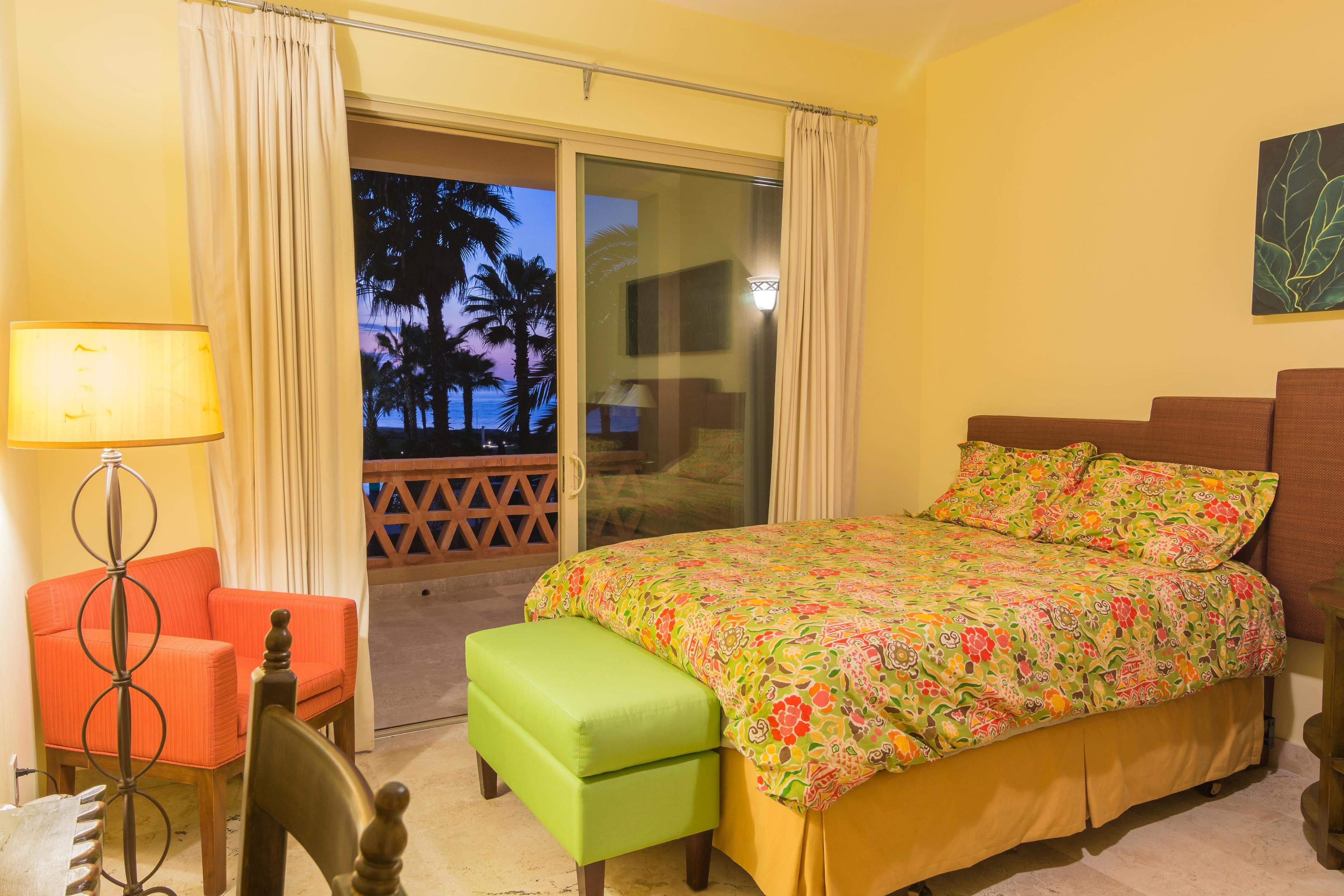 Habitación de hotel con cama, una silla, una lámpara y vistas a palmeras a través de una puerta corredera.
