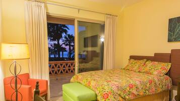 Habitación de hotel con cama, una silla, una lámpara y vistas a palmeras a través de una puerta corredera.