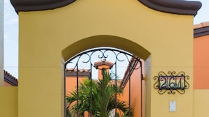 A yellow gate with a decorative metal design, a number plaque, and a view of a building with a tiled roof and potted plants. - Paraiso Del Mar Resort PDM V261 3 Bedroom Villa by Seaside La Paz (La Paz)