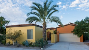 A two-story house with a yellow facade and an orange roof, featuring a garage and a well-maintained garden with various plants. - Paraiso Del Mar Resort PDM V261 3 Bedroom Villa by Seaside La Paz (La Paz)