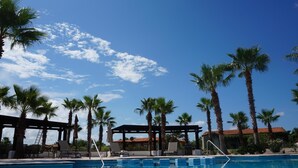 A pool area with lounge chairs, palm trees, and a building with a red roof. - Paraiso Del Mar Resort PDM V261 3 Bedroom Villa by Seaside La Paz (La Paz)