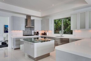 A modern kitchen with a central island, white cabinetry, and a large window providing a view of greenery. - Villa Venetian Star (Miami Beach)