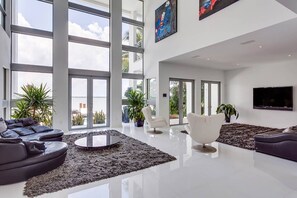 A spacious living room with large windows, a black leather sofa, a round coffee table, and a flat-screen TV mounted on the wall. - Villa Venetian Star (Miami Beach)