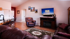 A living room with a leather sofa, a TV on a wooden stand, and a dining area with a kitchen in the background. - Gateway to Zion (La Verkin)