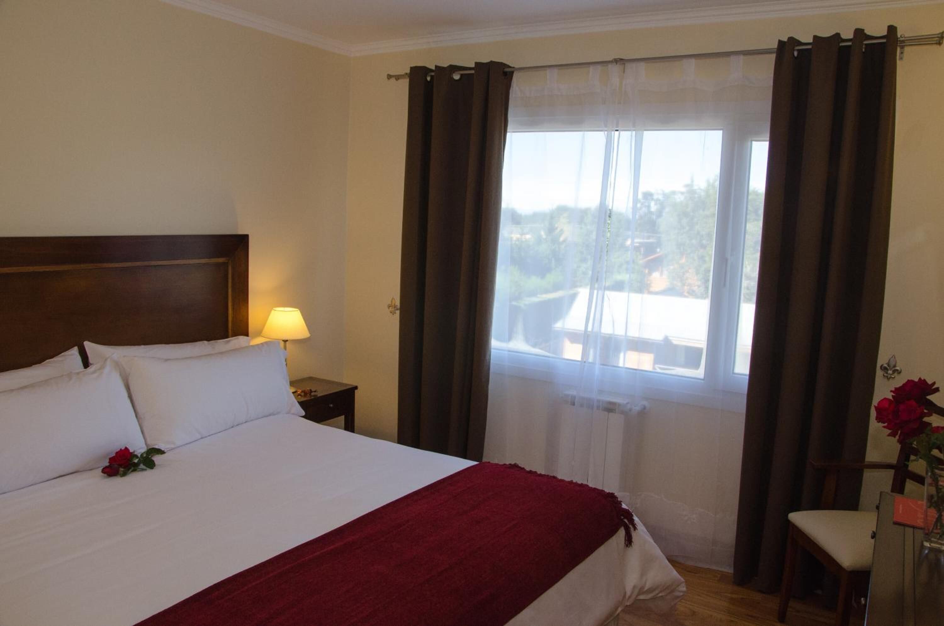 A hotel room with a bed, a nightstand, a lamp, a window with curtains, and a chair with a vase of flowers.