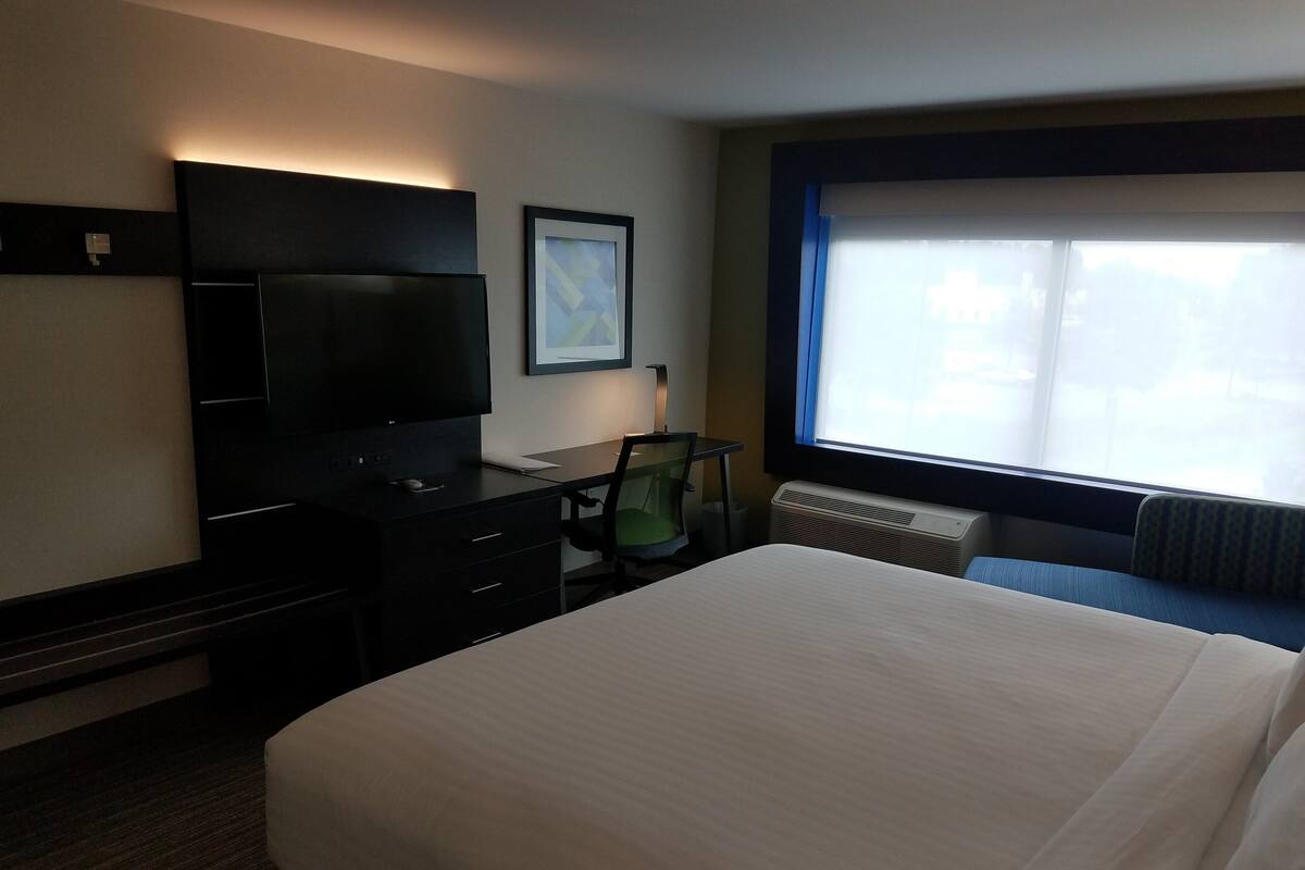 Habitación de hotel con cama, escritorio con ordenador, televisor, silla y ventana con persianas.