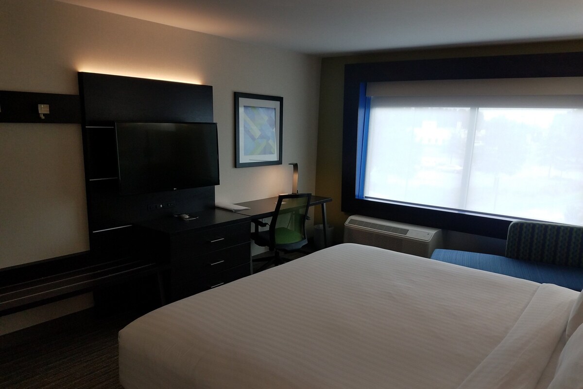 Habitación de hotel con cama, escritorio con ordenador, televisor, silla y ventana con persianas.