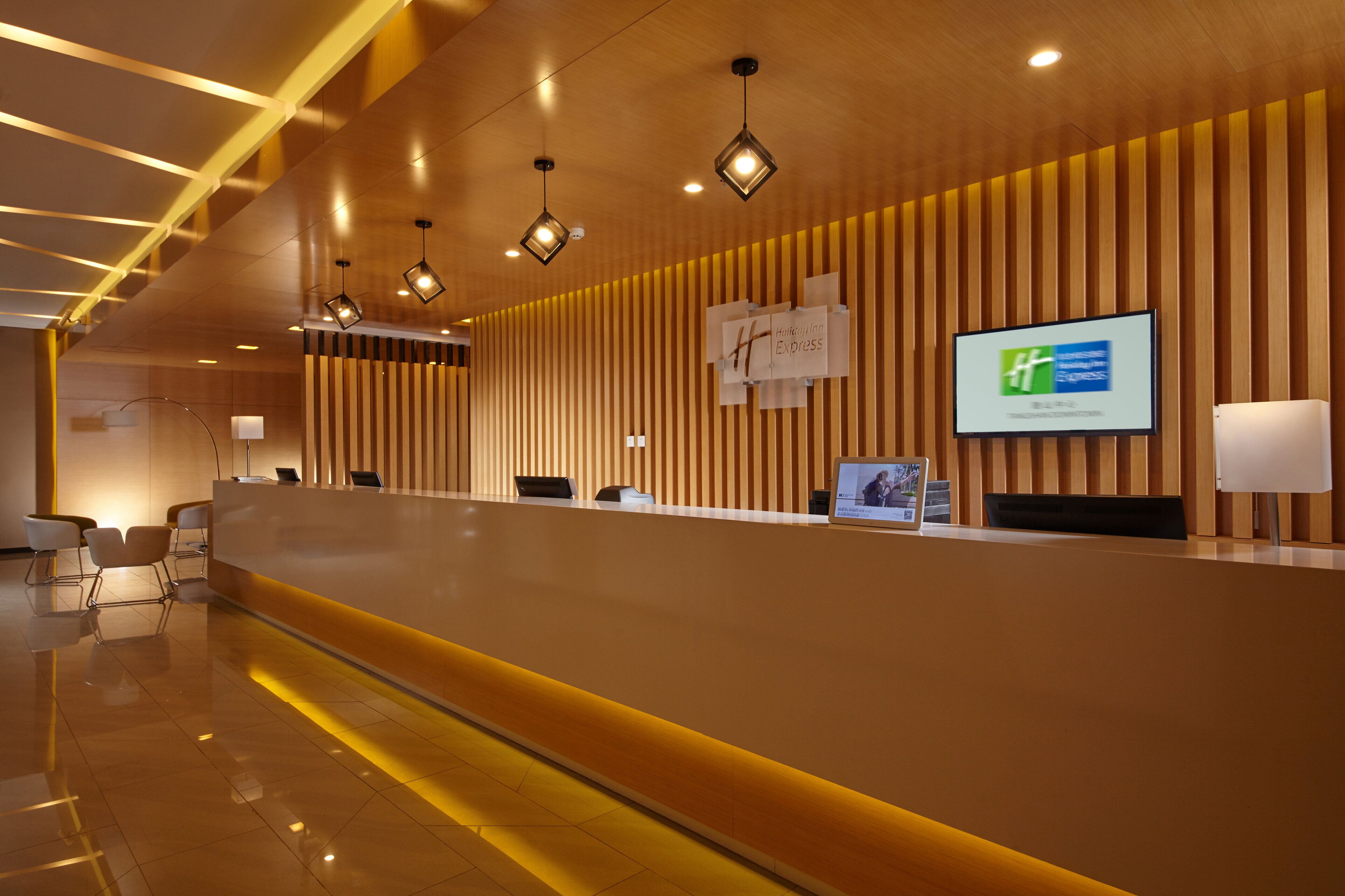 A modern reception area with a long reception desk, multiple chairs, and a large screen displaying the hotel name.