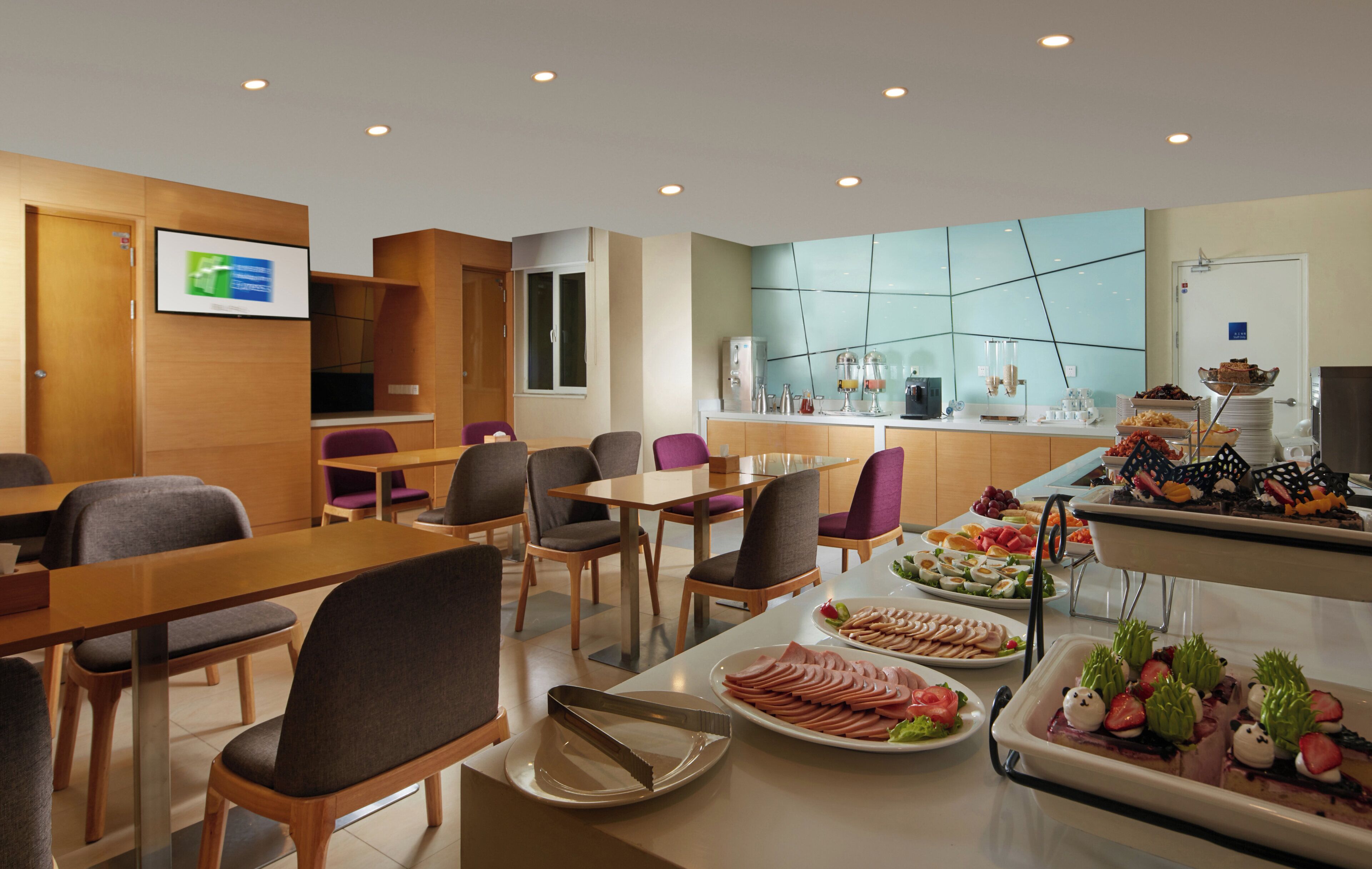 A hotel dining area with tables, chairs, a buffet with various food items, and a kitchen area in the background.