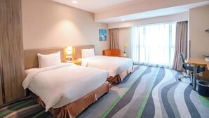 Quarto de hotel com duas camas, uma escrivaninha, uma cadeira e uma janela grande com cortinas.