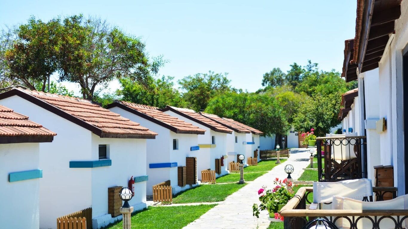 A row of white buildings with brown roofs, a paved pathway, and greenery in the background.